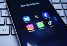 Na obrazovce telefonu je otevřená složka „Social Media“ se šesti ikonami aplikací: Bluesky, X (dříve Twitter), Facebook, Instagram, WhatsApp.