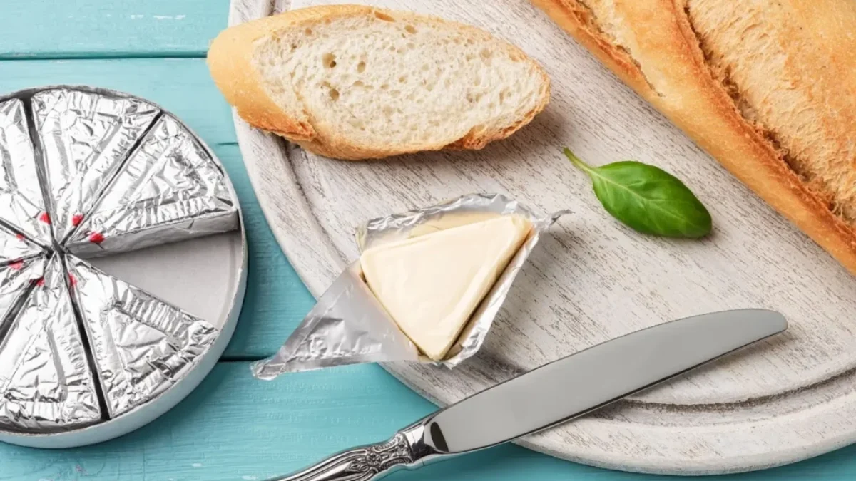Plátek bagety s rozbaleným trojúhelníčkem taveného sýra na dřevěném prkénku s nožem