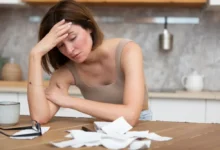 smutná, bezradná žena si drží hlavu v dlaních, na stole před sebou má účtenky